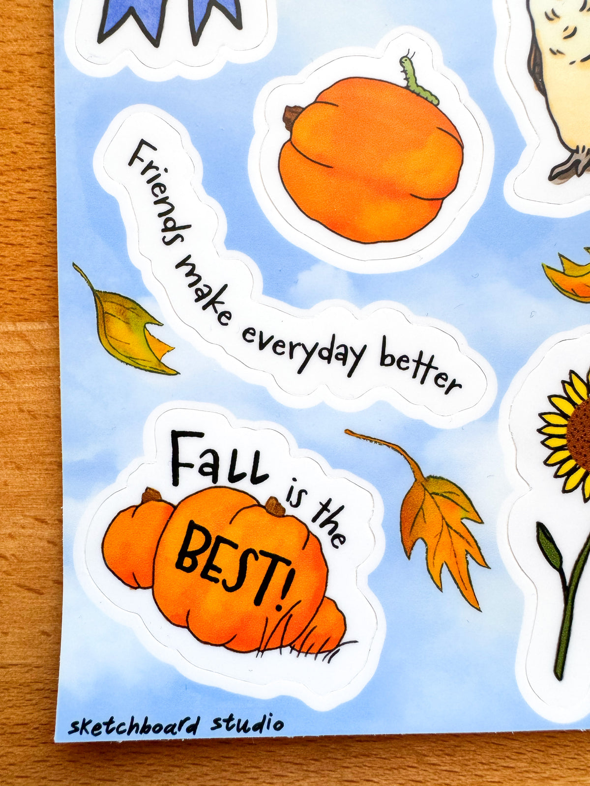 A close-up of a sheet of die-cut stickers featuring autumn illustrations and text, including pumpkins, leaves, a sunflower, and the phrases 'Friends make everyday better' and 'Fall is the BEST!'. The stickers are laid on a wooden surface.