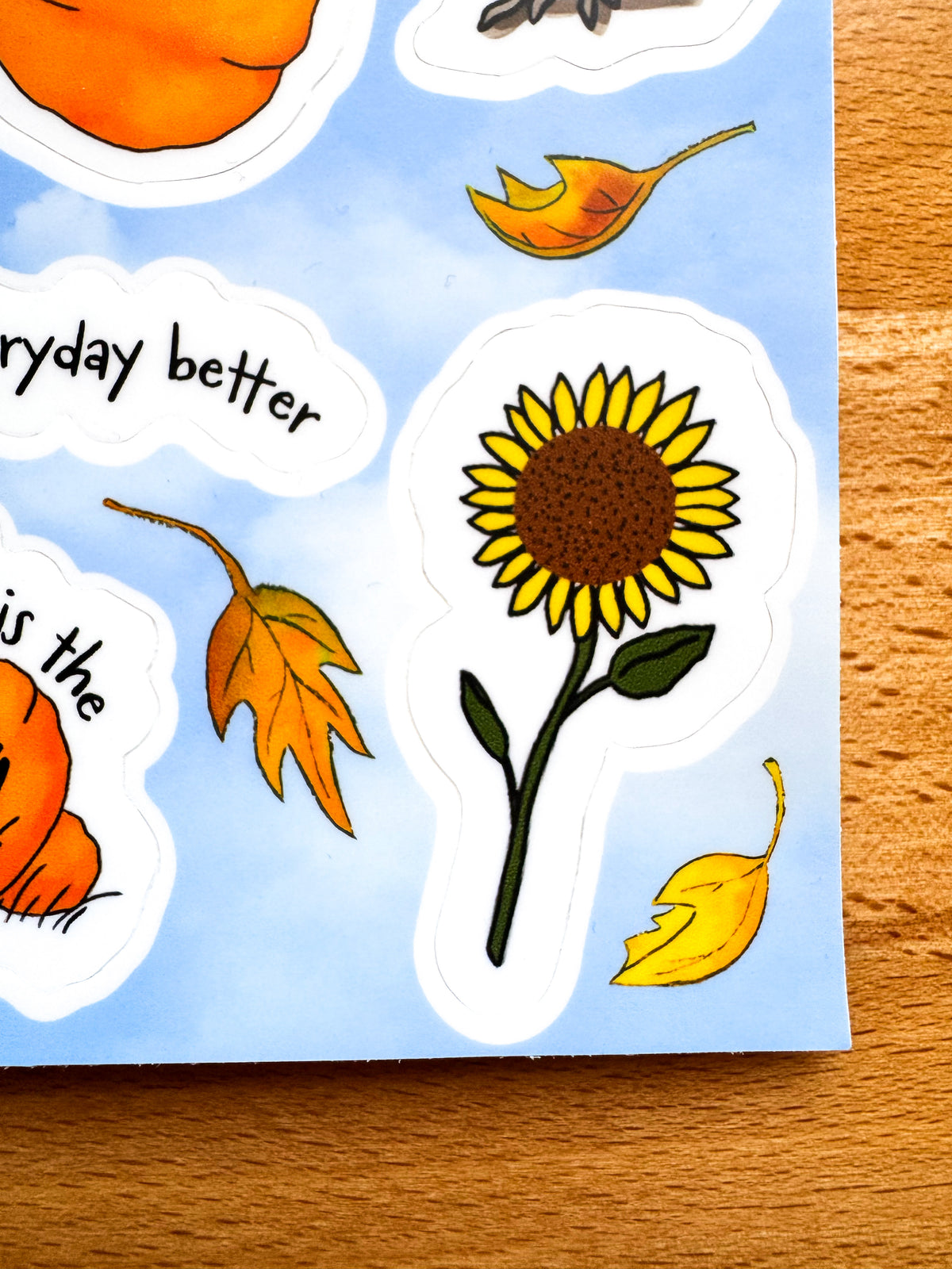 A close-up of a sticker sheet featuring autumn-themed designs, including a prominent die-cut sunflower sticker, orange pumpkins, yellow and orange leaves, and partial text reading 'everyday better' on a light blue background, placed on a wooden surface.