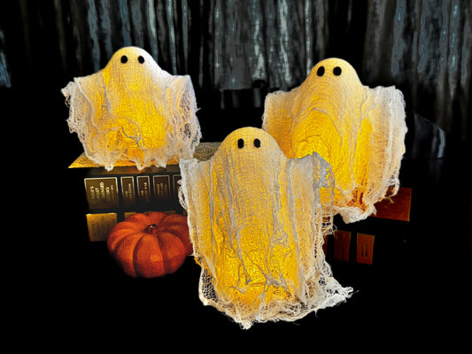 Three ghost-shaped Halloween decorations draped in white gauze, glowing with warm yellow light, displayed on a black surface with a small orange pumpkin and books in the background.