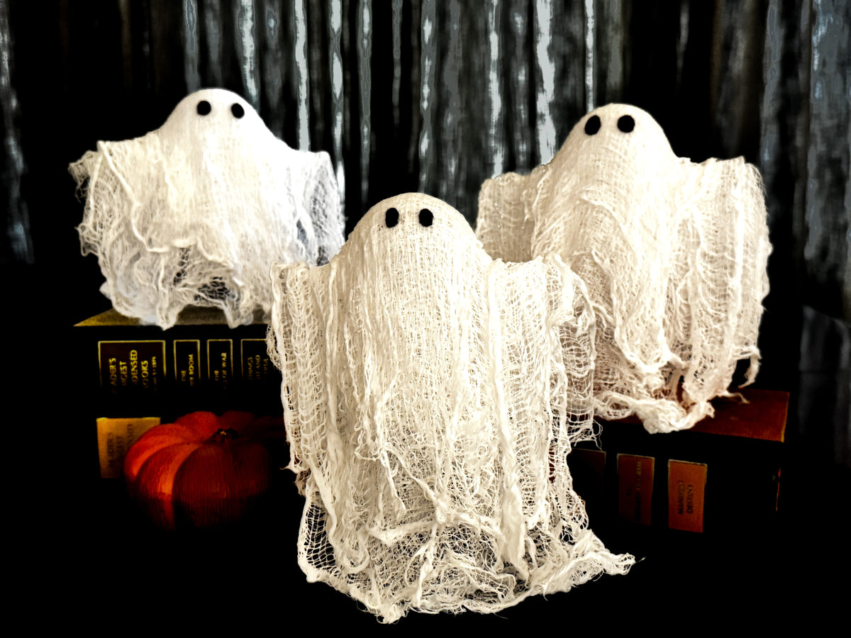 Three Halloween ghost decorations made of white gauze fabric with black bead eyes, displayed on top of old books with a small orange pumpkin on a black background.