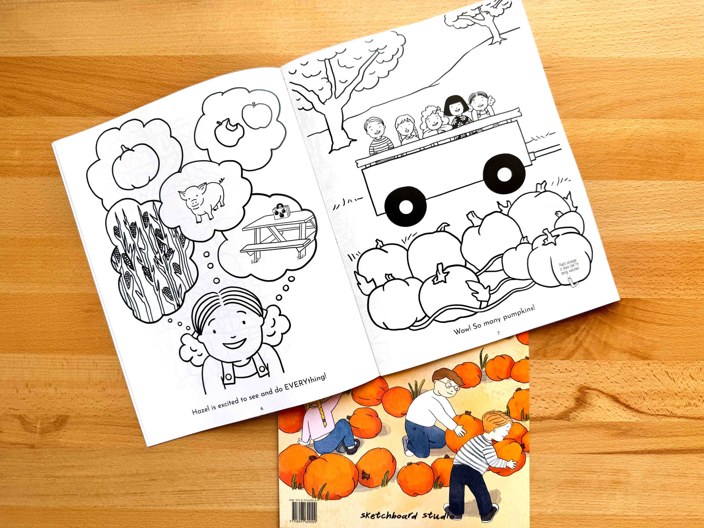 An open black-and-white children's coloring book lies on a wooden tabletop, with a second coloring book partially visible underneath. The top book shows illustrated scenes including pumpkins, a girl thinking about a pig and tractor, and a tractor loaded with children.