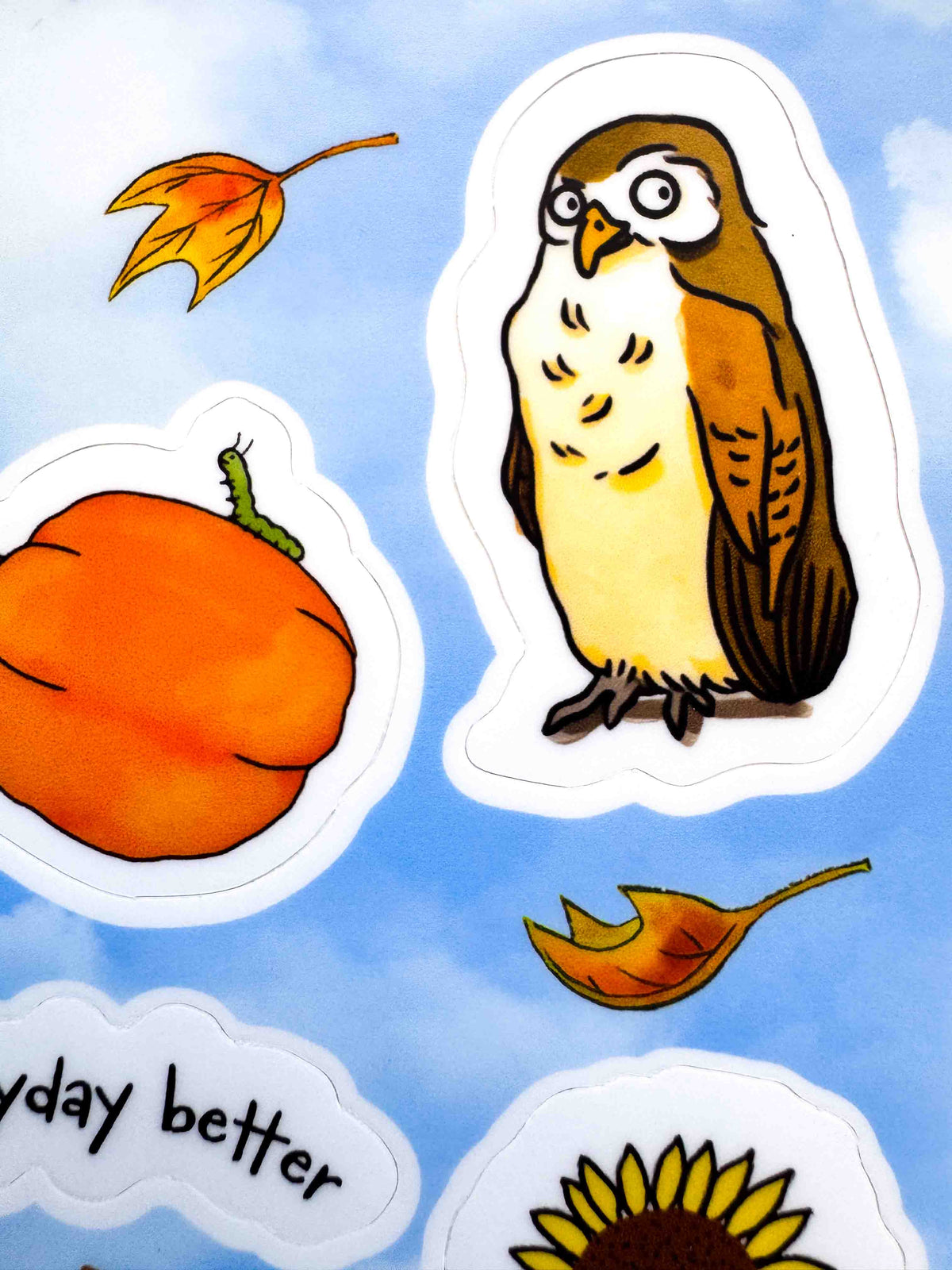 A close-up of autumn-themed die-cut stickers on a light blue sky background. The stickers include a brown and cream owl, an orange pumpkin, yellow and orange leaves, part of a sunflower, and a partial phrase reading 'day better'.