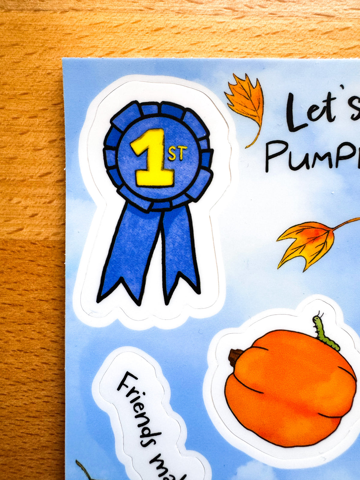 A close-up of a printed sticker sheet on a wooden surface, featuring a blue ribbon award sticker with yellow '1st' text, an orange pumpkin sticker, autumn leaves, and partial text stickers reading 'Let's Pump.